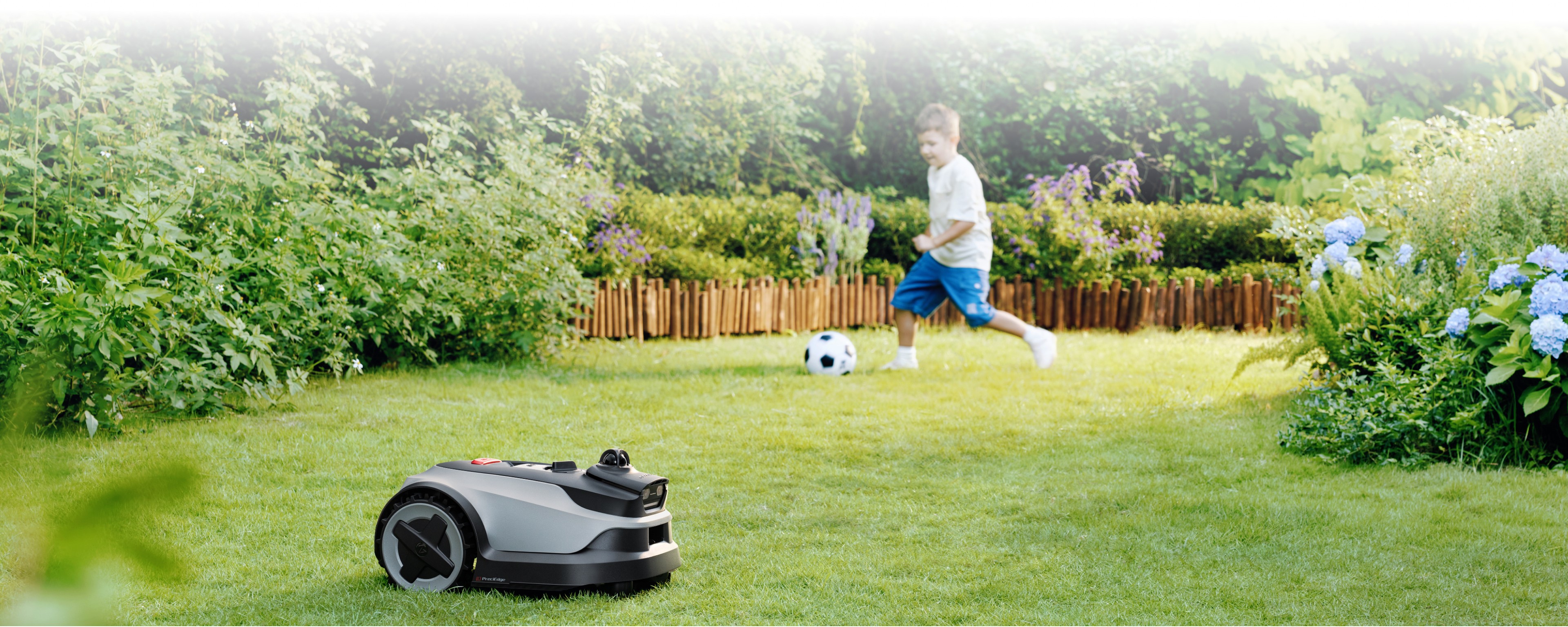 Family garden scene with boy playing soccer while robot mower mows in foreground